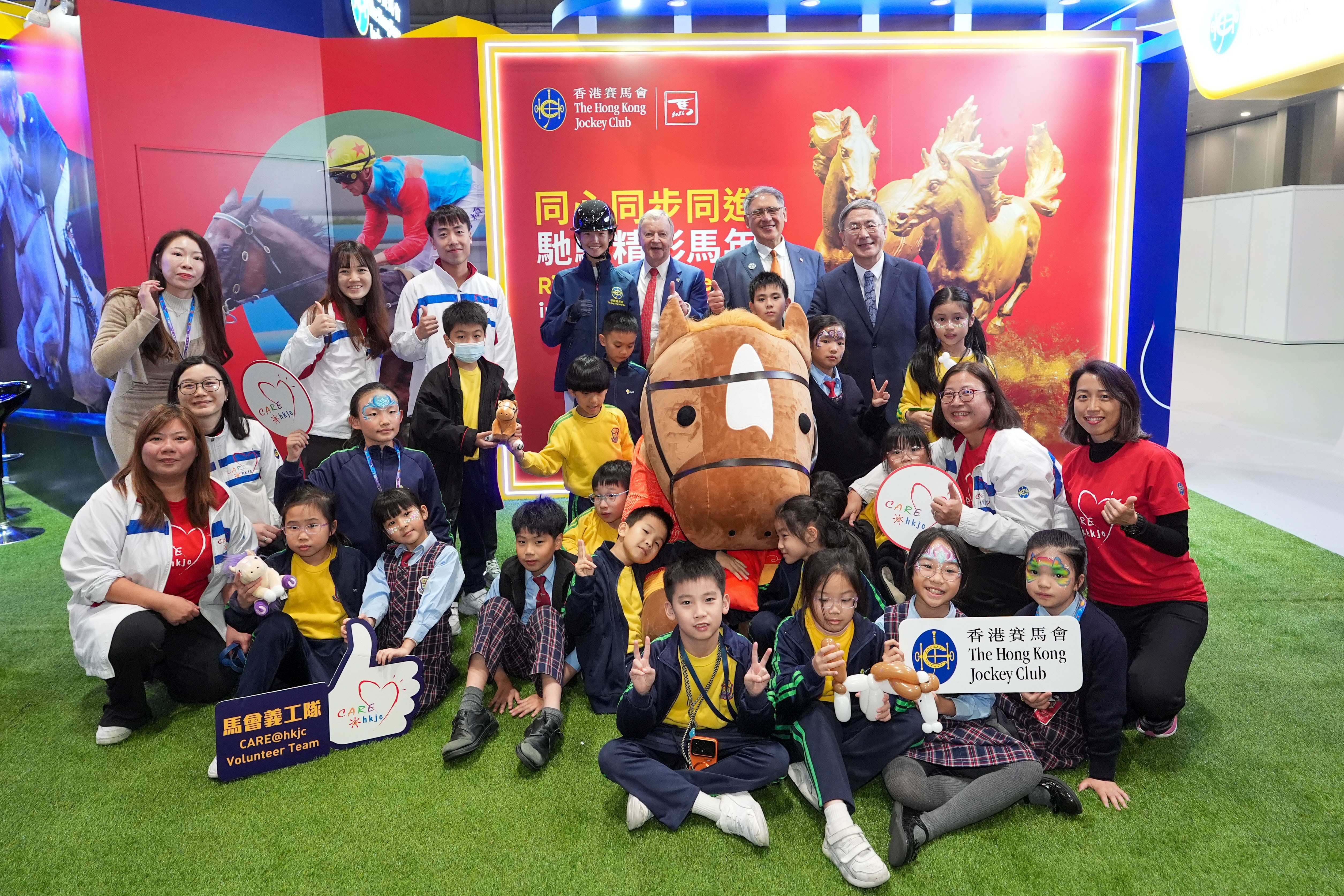 香港特別行政區政府政務司副司長卓永興 (後排右一)、香港賽馬會副主席兼浪琴香港國際馬術盛典主席及中國香港馬術總會會長黃嘉純 (後排右二)、香港賽馬會行政總裁應家柏 (後排右三)及馬會青少年馬術隊成員王嘉莉 (後排右四)，與馬會義工隊及進場觀賽的青年及基層團體，於馬會互動攤位合照